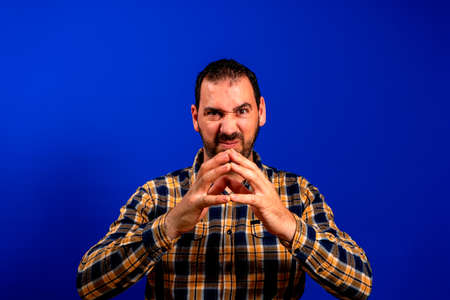 Bearded man in a yellow plaid shirt posing with folded hands in front of face in evil gesture...