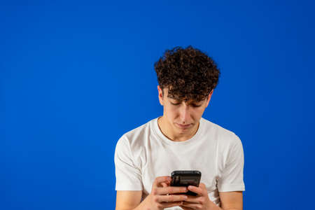 Handsome young man with curly hair using his smartphone isolated on blue studio background. Technology conceptの写真素材