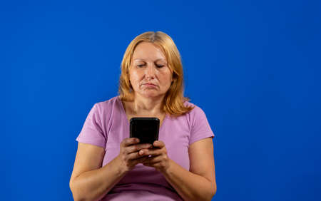 Pretty middle aged woman using her smartphone very serious isolated on blue studio background. Technology conceptの写真素材