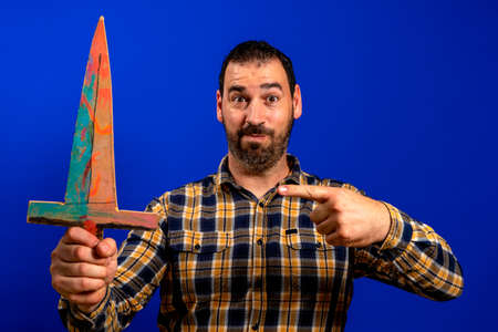 Man in a yellow plaid shirt scratching his back with a wooden sword isolated on blue studio background. Fun conceptの写真素材