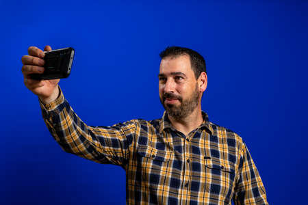 Bearded man in a yellow plaid shirt taking a selfie on blue studio background. Technology concept