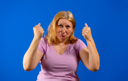 Middle age woman standing over blue isolated background angry and mad raising fists frustrated and furious while shouting with anger. Rage and aggressive concept.の写真素材