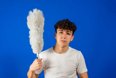 Caucasian young man posing funny with a cleaning duster on blue studio background. Cleaning conceptの写真素材