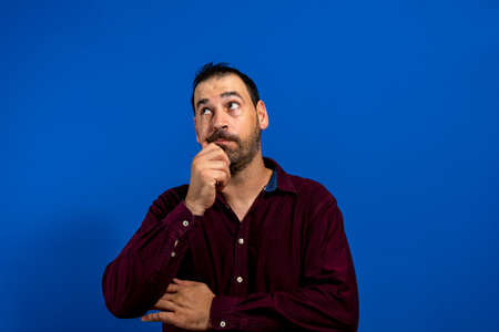Portrait of young thoughtful man with hand near face with pensative expression against blue...