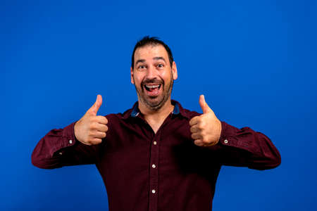 Portrait of cheerful man in basic clothing smiling and showing thumbs up at camera isolated over...