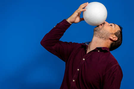 Close up of emotional caucasian man playing soccer hitting the ball with his mouth on isolated blue background. Soccer, sport, facial expression, human emotions, healthy lifestyle concept. Copyspaceの写真素材