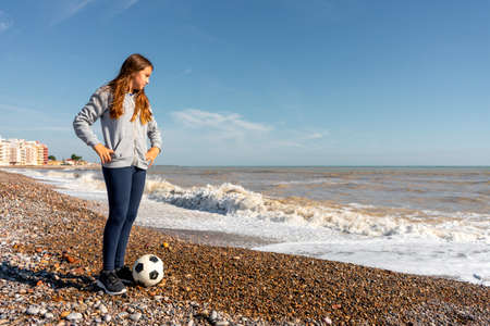 A beautiful emotional girl in a gray sweatshirt posing with her soccer ball while looking at the horizon on a pebble beach. Active winter holidays for kids.の写真素材