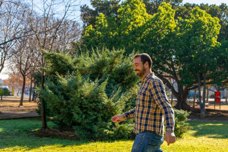 Hispanic man with a beard in his 40s wearing a plaid shirt and jeans walking smiling through the grass of a nice park.の写真素材
