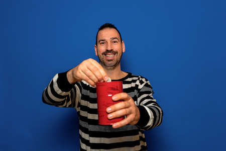 Smiling Latin man with a beard dressed in a striped sweater putting a euro coin in a metal piggy bank, trying to save so he can travel on his vacation. Isolated on blue studio background.の写真素材
