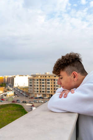 Young Caucasian man with curly hair dressed in a white sweatshirt looking at the city skyline serious and melancholic, I think about his future and what life has prepared for him.の写真素材