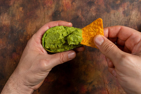 Man's hand dipping a triangle of corn in guacamole inside an empty avocado on a wooden background in brown tonesの写真素材