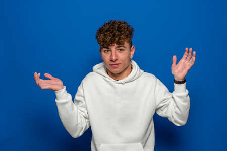 Young handsome brunette man with curly hair wearing white sweatshirt over blue background clueless and confused expression with arms and hands raised. doubt conceptの写真素材