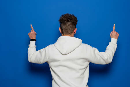 Portrait of a young man with curly hair from the back with index fingers pointing up isolated on blue studio backgroundの写真素材