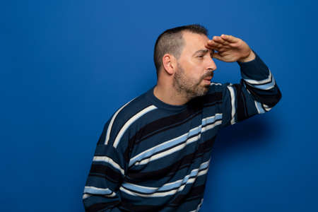Cheerful bearded young man in striped pullover posing isolated on blue background in studio. People lifestyle concept. Mock up copy space. Holding hand to forehead looking into the distanceの写真素材