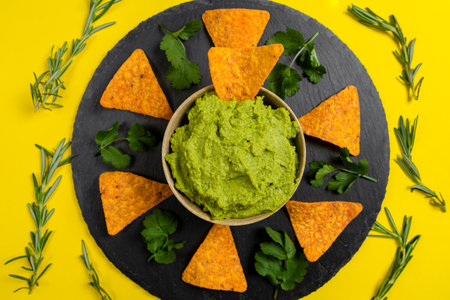 Composition of guacamole with aromatic herbs for decoration and nachos for consumption on a yellow backgroundの写真素材
