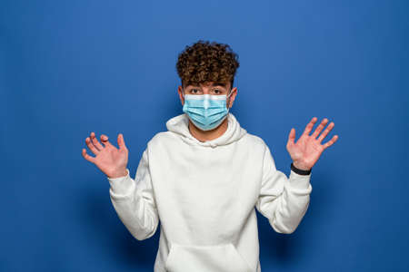 Shocked 20 year old man in white sweatshirt and sterile face mask safe from virus during quarantine raising hands showing palms isolated on blue background studio portrait.の写真素材
