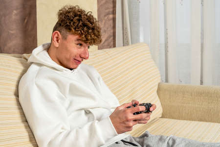 Thats my best round. Excited handsome young man with curly hair playing video games while he sitting on the couch at home.の写真素材