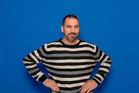Cheerful bearded man in striped pullover posing isolated on blue background, studio portrait. People lifestyle concept. Mock up copy space. Standing with your arms akimbo on your waist blinking.の写真素材
