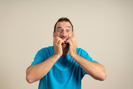 Studio portrait of scared and shocked dark-haired mature man biting his nails, staring wide-eyed at something awful, wearing casual shirt, standing over beige background, with copy space.の写真素材