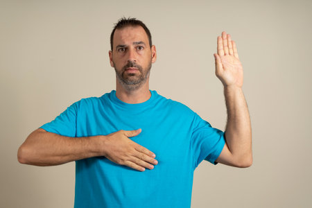 Bearded hispanic man in his 40s wearing blue t-shirt over beige isolated background swearing with hand on chest and open palm, making allegiance promise oath.の写真素材
