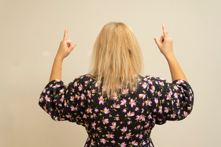 Latin blonde in a dark dress turned from behind pointing up with her index fingers, isolated over a beige background.の写真素材
