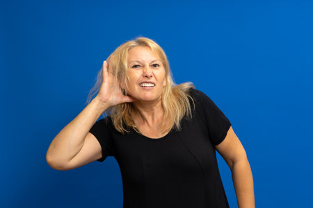 Caucasian blonde woman with her hand on her ear trying to hear something, snoops and gossips about any rumors. Isolated on blue studio background.の写真素材