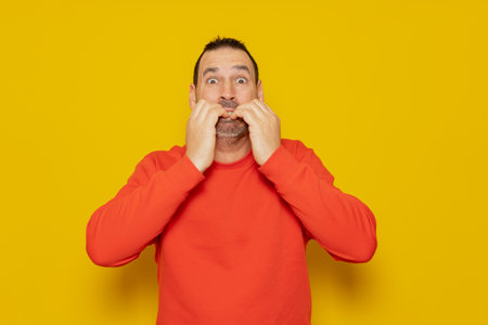 Studio portrait of scared and shocked dark-haired mature man biting his nails, wide-eyed looking at something horrible, wearing red jumper, standing over yellow background, copy spaceの写真素材