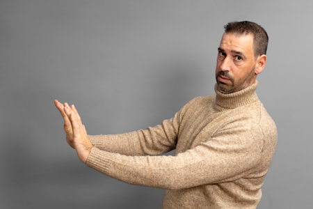 Side view of serious man making stop gesture showing palm, conflict ban warning about danger, stop bullying, wears beige turtleneck sweater. Indoor studio shot isolated on gray backgroundの写真素材