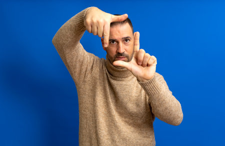 Handsome bearded happy male looks through a frame formed by his hands. An attractive man makes a frame with his fingers, isolated on a blue background with copy space for text. People concept.の写真素材