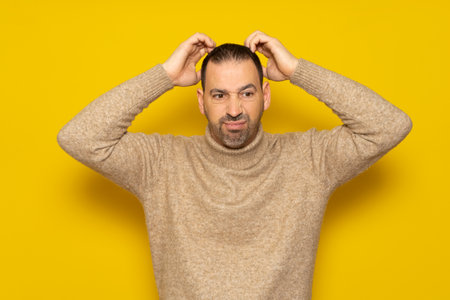Bearded Hispanic man wearing a beige turtleneck scratching his head energetically while trying to solve a problem or doubt, isolated over yellow background.の写真素材