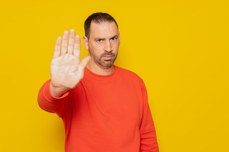 Hispanic man with beard in red sweater making stop gesture while looking angrily at camera, isolated...