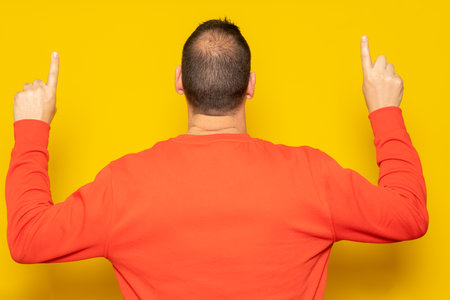 Hispanic man in red sweatshirt pointing up with his index fingers with his back to the camera, isolated on yellow background.の写真素材