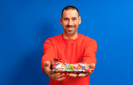 Bearded Hispanic man in his 40s wearing a red sweater delivering a birthday present to his beloved, he smiles with satisfaction as his partner has been pleasantly surprised. Isolated blue background.の写真素材