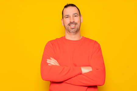 Portrait of a smart, cheerful and attractive bearded Hispanic man in his 40s, arms crossed, isolated over yellow colored backgroundの写真素材