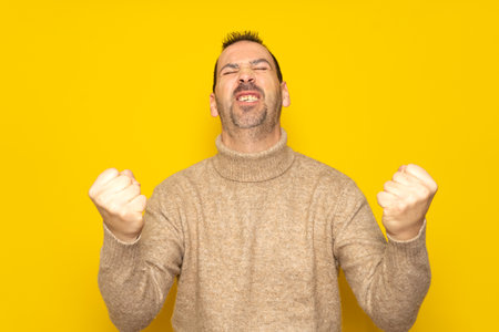 Bearded Hispanic man in his 40s wearing a beige turtleneck energetically celebrating a big win with raised fists, isolated on yellow studio backgroundの写真素材