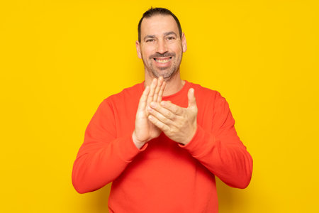Bearded hispanic man in his 40s wearing a red jumper clapping hands happy and cheerful, smiling proud hands together. Isolated on yellow studio backgroundの写真素材