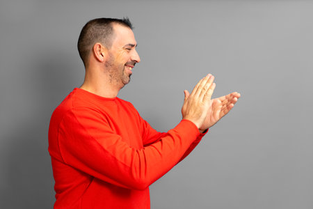 Bearded hispanic man in profile wearing a red jumper feeling happy, smiling and clapping with grateful emphasis for the great show he just witnessed. Isolated on gray studio background.の写真素材