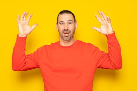 Bearded Hispanic man in his 40s wearing a red jumper tremendously surprised by an unexpected visitor, raises his arms excited. Isolated on yellow studio background.の写真素材