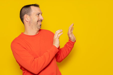 Bearded hispanic man in his 40s wearing a red jumper standing to the side using his hands for protection totally embarrassed and embarrassed. Isolated on yellow backgroundの写真素材