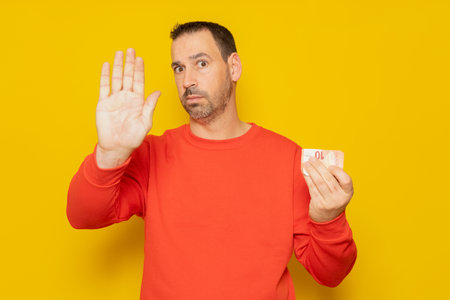 Bearded hispanic man in his 40s wearing a red jumper holding a 10 euro banknote and making the stop sign isolated over yellow backgroundの写真素材