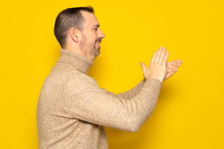 Bearded hispanic man in profile wearing brown turtleneck feeling happy, smiling and clapping hands with emphasis thankful for the great show he just witnessed. Isolated on yellow studio backgroundの写真素材