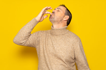 Bearded Hispanic man wearing a beige turtleneck smacking his lips and licking his fingers after devouring an exquisite delicacy, isolated on yellow studio backgroundの写真素材