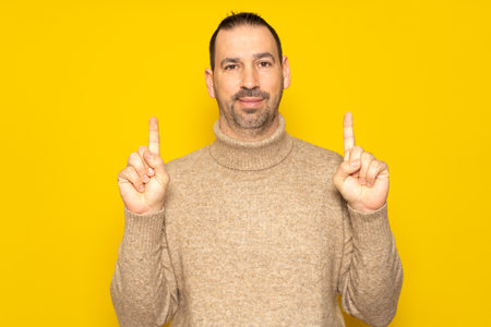 Bearded Hispanic man in his 40s wearing a beige sweater in a friendly attitude pointing up with his index fingers, isolated on yellow studio backgroundの写真素材