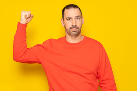 Bearded hispanic man wearing a red jersey flexing his biceps muscle showing his pitiful state of shape, isolated over yellow backgroundの写真素材