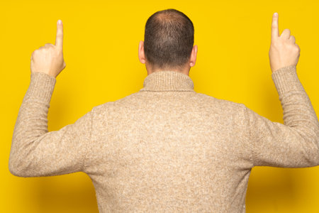 Hispanic man in casual clothing posing backwards pointing up with his index fingers, isolated on yellow studio background.の写真素材