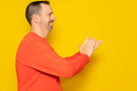 Bearded hispanic man in profile wearing a red jumper feeling happy, smiling and clapping with grateful emphasis for the great show he just witnessed. Isolated on yellow studio background.の写真素材