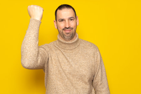 Bearded Hispanic man in his 40s wearing a beige turtleneck raising his fist while smiling with a victorious expression, isolated over yellow studio background.の写真素材