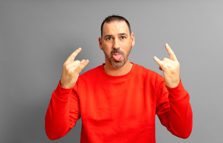 Latino man with a beard in his 40s wearing a red sweater making the rock gesture while sticking out his tongue in a crazy attitude, isolated on yellow studio background.の写真素材