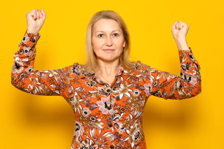 Strong and powerful caucasian woman, raises her arms and shows biceps, wears a patterned shirt, pattern on a yellow background. look at my muscles.の写真素材
