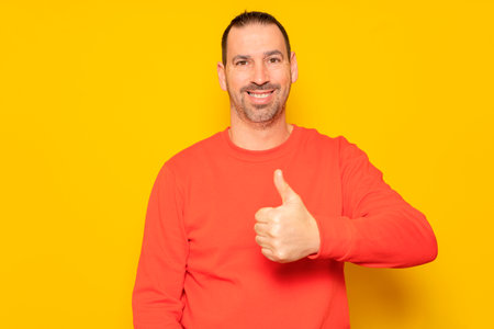 Bearded Hispanic man in his 40s wearing a red jumper proudly raising his thumb up while looking and smiling at the camera, isolated on yellow studio background.の写真素材